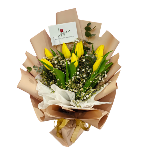 Bright yellow tulips bouquet with white gypsophila, yellow limonium, and eucalyptus foliage, wrapped in soft floral paper with ribbon.
